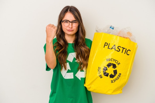 Junge kaukasische Frau, die einen vollen Kunststoff recycelt, der auf weißem Hintergrund isoliert ist und die Faust zur Kamera zeigt, aggressiver Gesichtsausdruck.