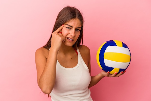 Junge kaukasische Frau, die einen Volleyballball einzeln auf rosafarbenem Hintergrund hält und eine Enttäuschungsgeste mit dem Zeigefinger zeigt.
