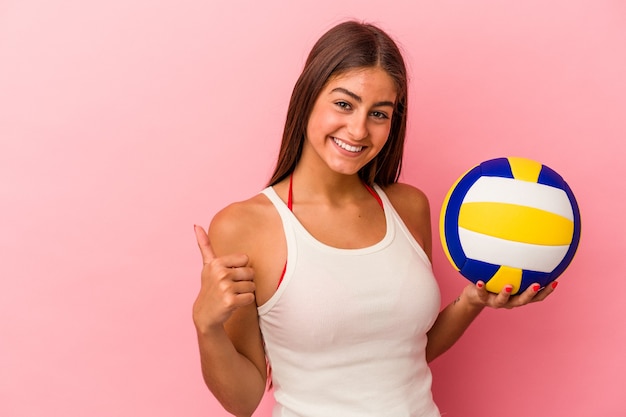 Junge kaukasische Frau, die einen Volleyballball hält, der auf rosa Hintergrund lokalisiert wird, lächelt und Daumen hochhebt