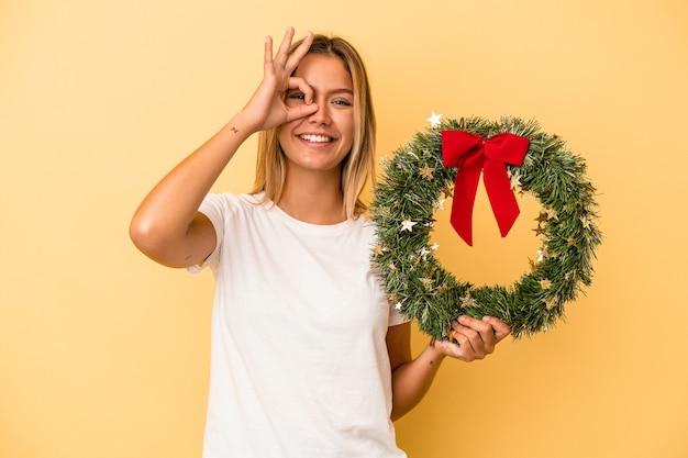 Junge kaukasische Frau, die einen Weihnachtskranz lokalisiert auf gelbem Hintergrund hält, aufgeregt, ok Geste auf Auge zu halten.