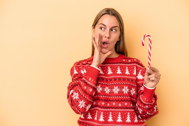 Junge kaukasische Frau, die einen Weihnachtsstock isoliert auf gelbem Hintergrund hält, sagt eine geheime heiße Bremsnachricht und schaut beiseite