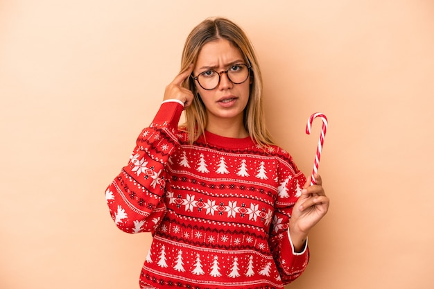 Junge kaukasische Frau, die einen Weihnachtsstock lokalisiert auf beigem Hintergrund hält, der eine Enttäuschungsgeste mit dem Zeigefinger zeigt.