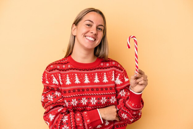 Junge kaukasische Frau, die einen Weihnachtsstock lokalisiert auf gelbem Hintergrund hält, lacht und Spaß hat.