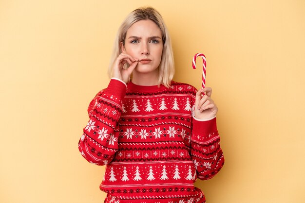 Foto junge kaukasische frau, die einen weihnachtsstock lokalisiert auf gelbem hintergrund mit den fingern auf den lippen hält, die ein geheimnis halten.