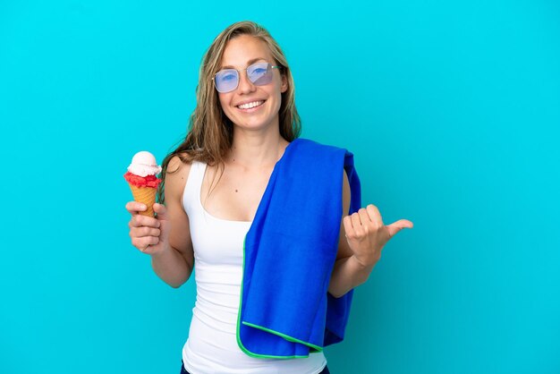 Junge kaukasische Frau, die Eis hält und ein Strandtuch trägt, das auf blauem Hintergrund isoliert ist und zur Seite zeigt, um ein Produkt zu präsentieren