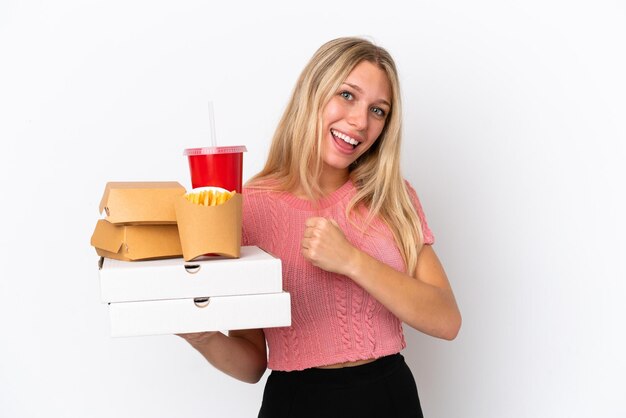 Junge kaukasische Frau, die fettes Essen isoliert auf blauem Hintergrund hält und einen Sieg feiert