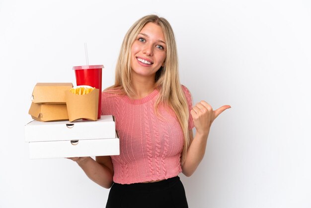 Junge kaukasische Frau, die fettes Essen isoliert auf blauem Hintergrund hält und zur Seite zeigt, um ein Produkt zu präsentieren