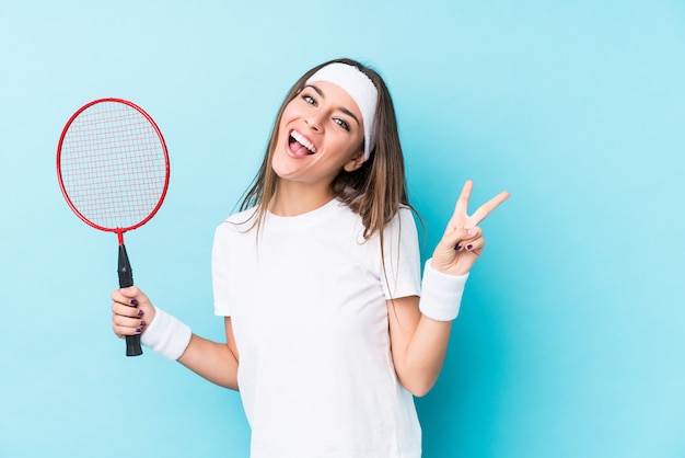 Junge kaukasische Frau, die freudig und sorglos Badminton spielt und ein Friedenssymbol mit den Fingern zeigt.