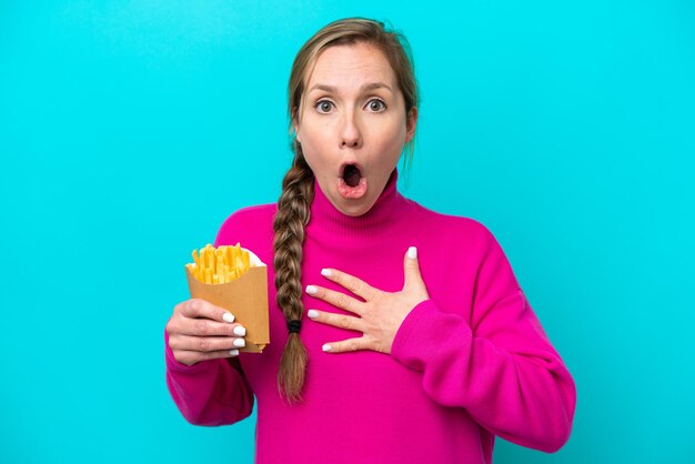 Junge kaukasische Frau, die gebratene Chips isoliert auf blauem Hintergrund hält, überrascht und schockiert, während sie nach rechts schaut