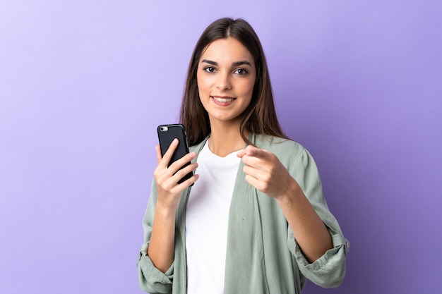 Junge kaukasische Frau, die Handy auf lila Zeigefinger auf Sie mit einem selbstbewussten Ausdruck verwendet