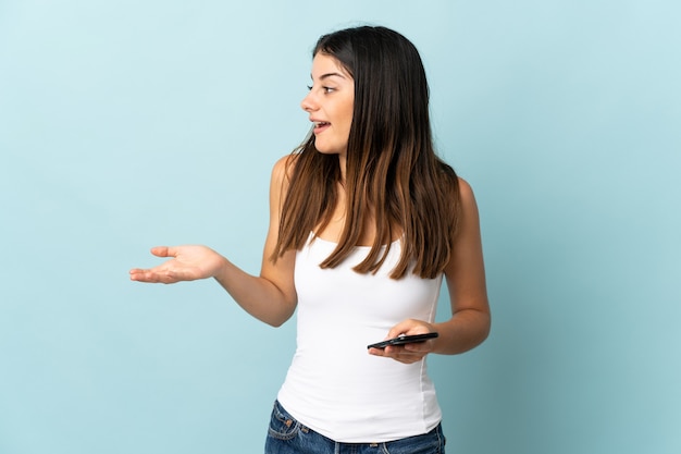 Junge kaukasische Frau, die Handy lokalisiert auf Blau mit Überraschungsausdruck beim Betrachten der Seite verwendet