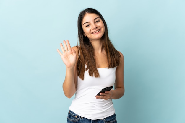 Junge kaukasische Frau, die Handy lokalisiert auf blau salutierend mit Hand mit glücklichem Ausdruck verwendet