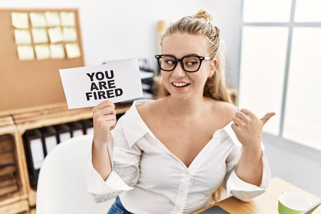 Junge kaukasische Frau, die im Büro ein „Sie sind gefeuert“-Banner hochhält und mit dem Daumen zur Seite zeigt, glücklich lächelnd und mit offenem Mund