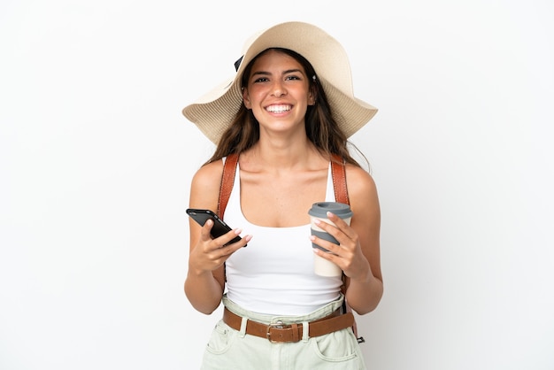 Junge kaukasische Frau, die in den Sommerferien eine Pamela trägt, isoliert auf weißem Hintergrund, die Kaffee zum Mitnehmen und ein Handy hält