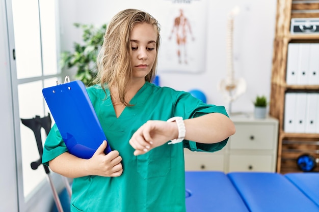 Junge kaukasische Frau, die in einer Schmerzklinik arbeitet und die Zeit auf der Armbanduhr überprüft, entspannt und selbstbewusst