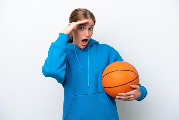 Junge kaukasische Frau, die isoliert auf weißem Hintergrund Basketball spielt und dabei eine Überraschungsgeste macht, während sie zur Seite schaut