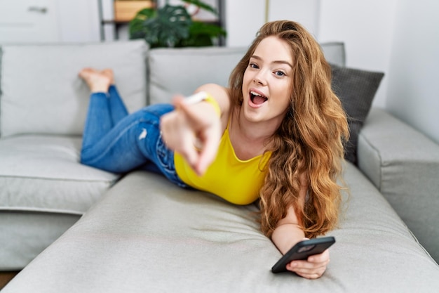 Junge kaukasische Frau, die mit dem Smartphone auf dem Sofa liegt und mit dem Finger nach vorne zeigt, überrascht mit offenem Mund, etwas auf der Vorderseite