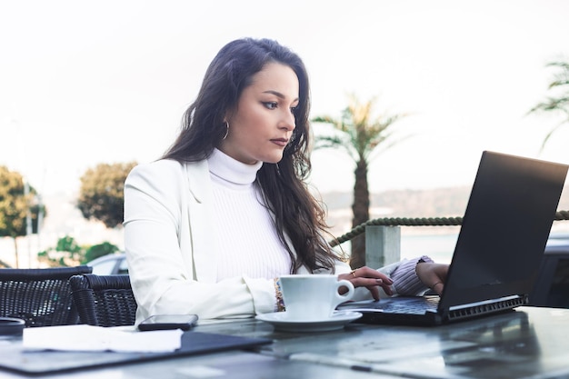 Junge kaukasische Frau, die mit einem Laptop arbeitet, während sie eine Kaffeepause auf einer Terrasse neben dem Strand macht