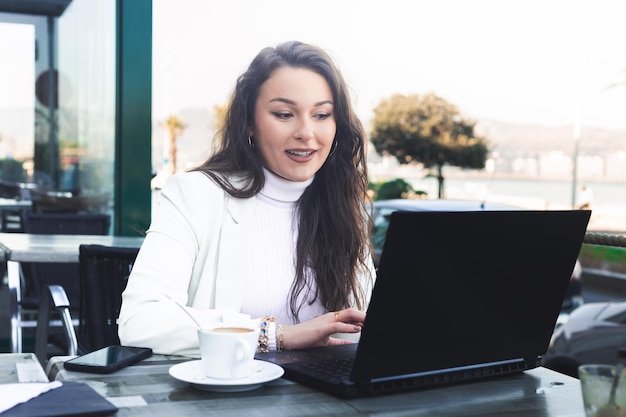 Junge kaukasische Frau, die mit einem Laptop arbeitet, während sie eine Kaffeepause auf einer Terrasse neben dem Strand macht