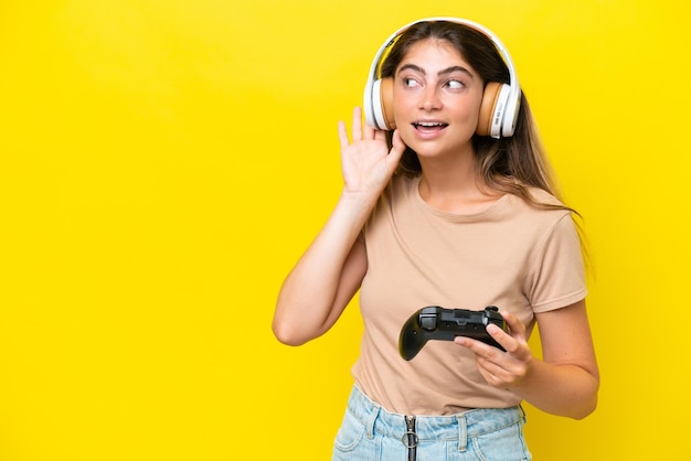 Junge kaukasische Frau, die mit einem Videospiel-Controller spielt, der auf gelbem Hintergrund isoliert ist und etwas hört, indem sie die Hand auf das Ohr legt