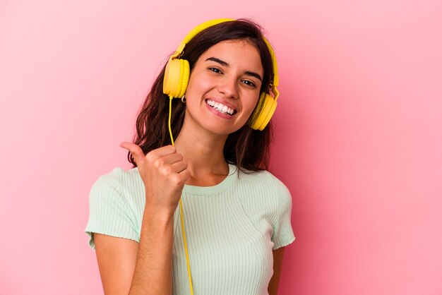 Junge kaukasische Frau, die Musik hört, die auf rosafarbenem Hintergrund lokalisiert wird, lächelt und hebt Daumen hoch