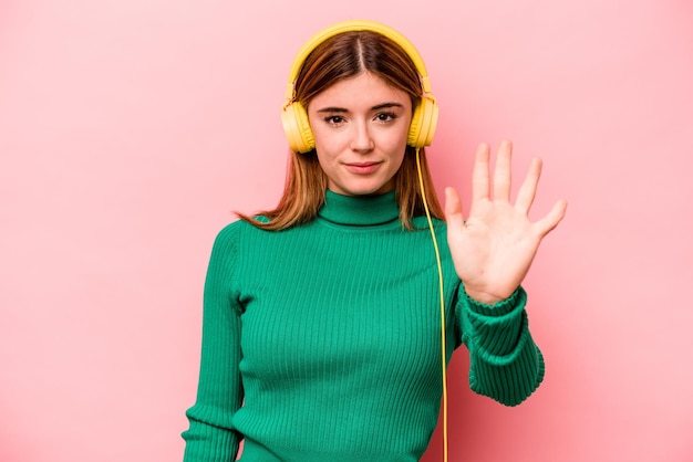 Junge kaukasische Frau, die Musik hört, isoliert auf rosa Hintergrund, lächelt fröhlich und zeigt Nummer fünf mit den Fingern