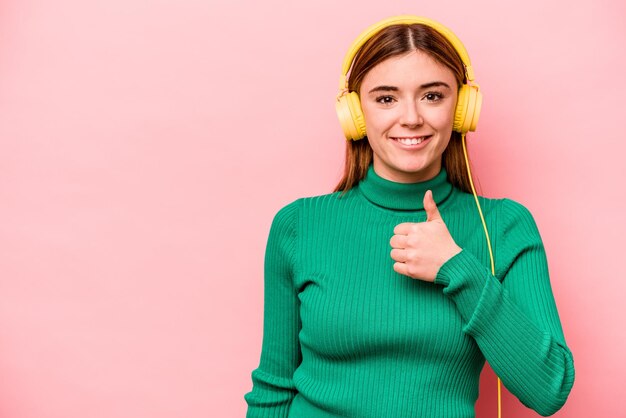 Junge kaukasische Frau, die Musik hört, isoliert auf rosa Hintergrund, lächelt und hebt den Daumen nach oben