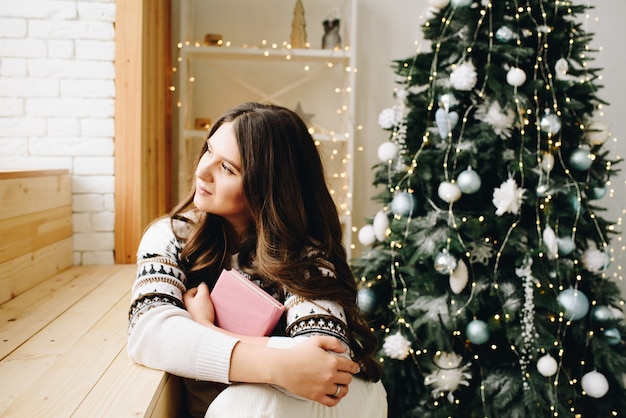 Junge kaukasische Frau, die neben reizendem geschmücktem Weihnachtsbaum sitzt, der ein Buch hält und träumt