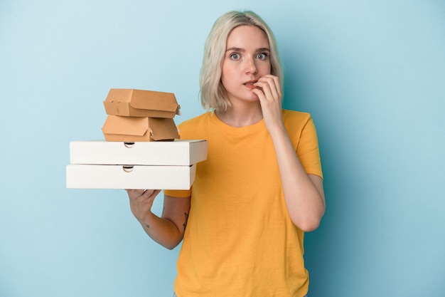 Junge kaukasische Frau, die Pizza und Burger einzeln auf blauem Hintergrund hält und Fingernägel beißt, nervös und sehr ängstlich.