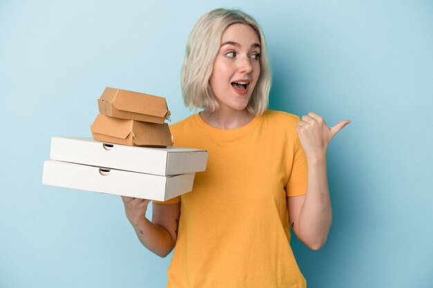 Junge kaukasische Frau, die Pizza und Burger einzeln auf blauem Hintergrund hält, zeigt mit dem Daumenfinger weg, lacht und sorglos.