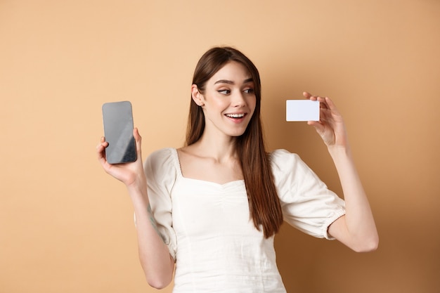 Junge kaukasische Frau, die Plastikkreditkarte mit erfreutem Lächeln zeigt, demonstrieren leeren Handybildschirm, der auf beigem Hintergrund steht.