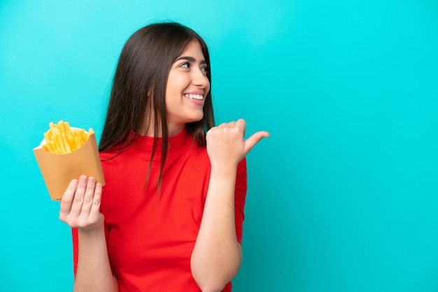 Junge kaukasische Frau, die Pommes frites fängt, isoliert auf blauem Hintergrund, der zur Seite zeigt, um ein Produkt zu präsentieren