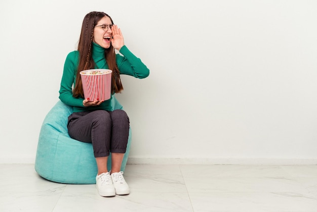 Junge kaukasische Frau, die Popcorn auf einem Puff isst, isoliert auf weißem Hintergrund, schreit und die Handfläche in der Nähe des geöffneten Mundes hält