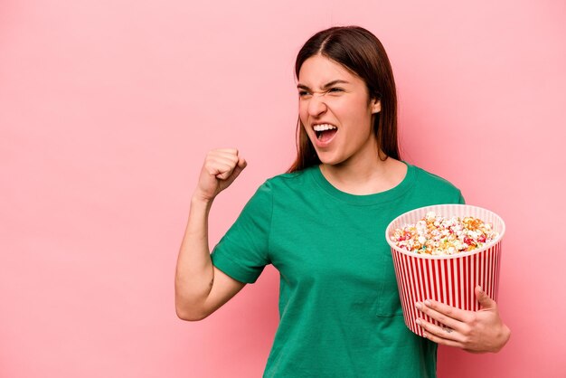 Junge kaukasische Frau, die Popcorn isoliert auf rosafarbenem Hintergrund hält und die Faust nach einem Siegerkonzept erhebt