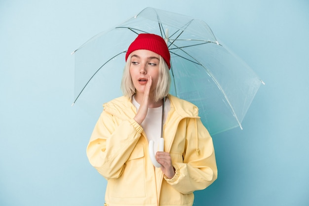 Junge kaukasische Frau, die Regenschirm isoliert auf blauem Hintergrund hält, sagt eine geheime heiße Bremsnachricht und schaut beiseite