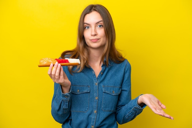 Junge kaukasische Frau, die Sashimi isoliert auf gelbem Hintergrund hält und Zweifel macht, während sie die Schultern anhebt