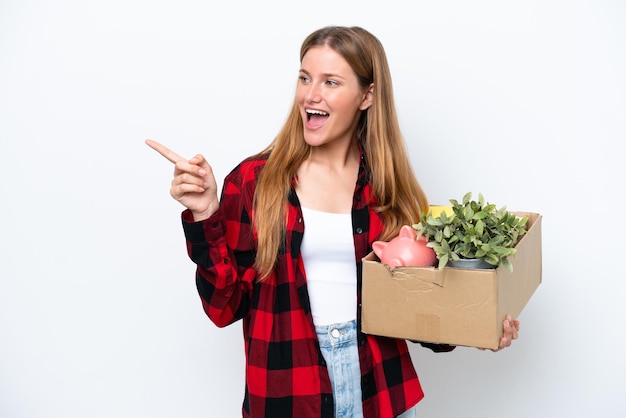 Junge kaukasische Frau, die sich bewegt, während sie eine Kiste voller Dinge aufhebt, die auf weißem Hintergrund isoliert sind, mit dem Finger zur Seite zeigt und ein Produkt präsentiert