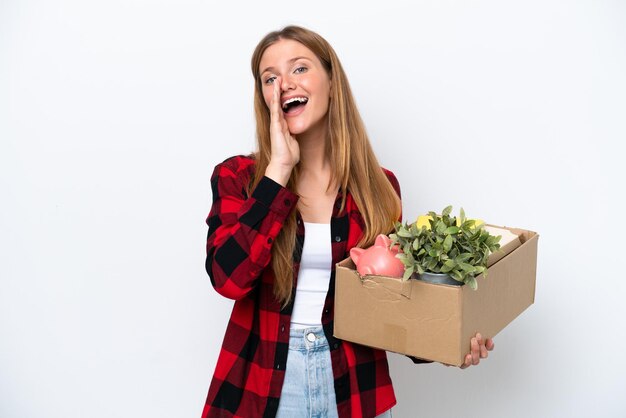 Junge kaukasische Frau, die sich bewegt, während sie eine Kiste voller Dinge aufhebt, die auf weißem Hintergrund isoliert sind und mit weit geöffnetem Mund schreien