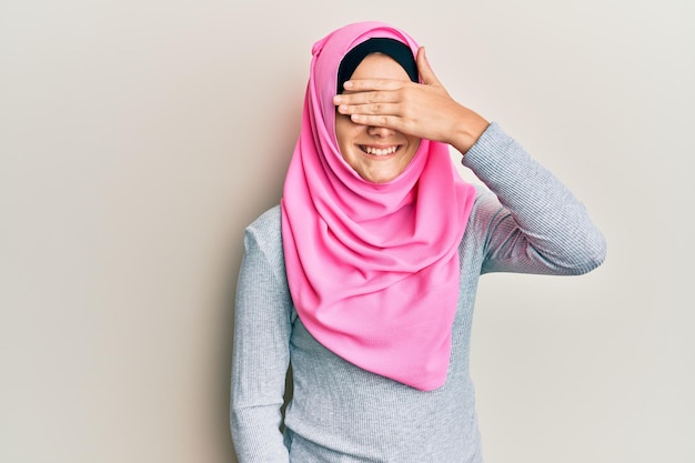 Junge kaukasische Frau, die traditionellen islamischen Hijab-Schal trägt, lächelt und lacht mit der Hand auf dem Gesicht, die die Augen bedeckt, für ein überraschendes blindes Konzept