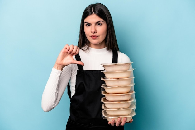 Junge kaukasische Frau, die Tupperware isoliert auf blauem Hintergrund hält und eine Abneigungsgeste mit dem Daumen nach unten zeigt Uneinigkeitskonzept