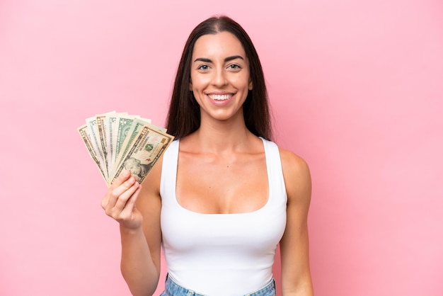 Junge kaukasische Frau, die viel Geld isoliert auf rosafarbenem Hintergrund nimmt und viel lächelt