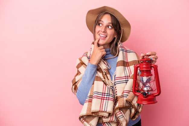 Junge kaukasische Frau, die Vintage-Laterne einzeln auf rosafarbenem Hintergrund hält, entspannte sich beim Nachdenken über etwas, das einen Kopienraum betrachtet.