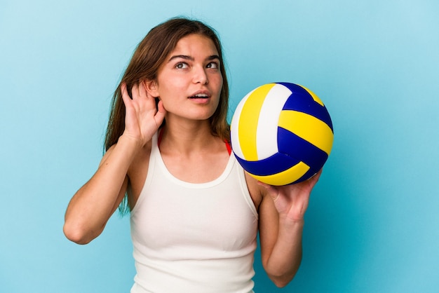 Junge kaukasische Frau, die Volleyball auf blauem Hintergrund isoliert spielt und versucht, einen Klatsch zu hören.