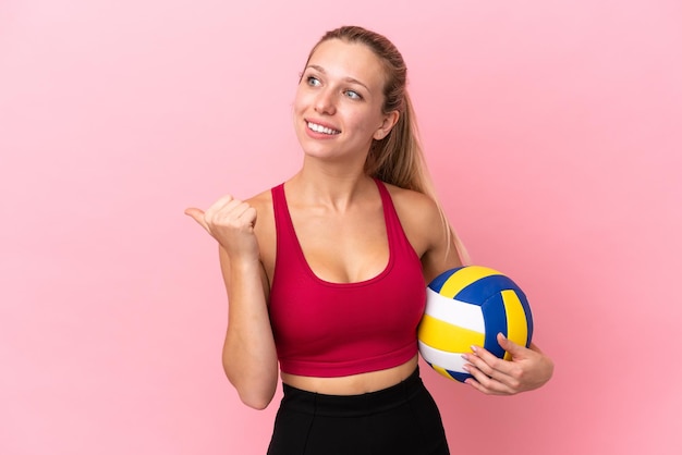 Junge kaukasische Frau, die Volleyball spielt, isoliert auf rosafarbenem Hintergrund, der zur Seite zeigt, um ein Produkt zu präsentieren