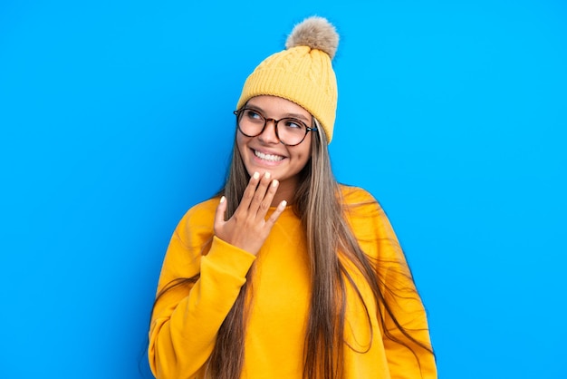 Junge kaukasische Frau, die Winterkleidung trägt, die auf blauem Hintergrund isoliert aufschaut, während sie lächelt