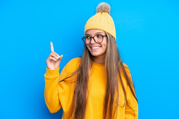 Junge kaukasische Frau, die Winterkleidung trägt, isoliert auf blauem Hintergrund, die auf eine großartige Idee zeigt