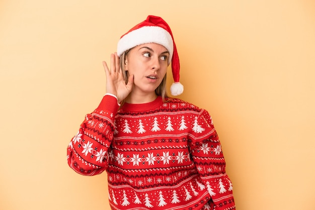 Junge kaukasische Frau feiert Weihnachten isoliert auf gelbem Hintergrund und versucht, einen Klatsch zu hören.