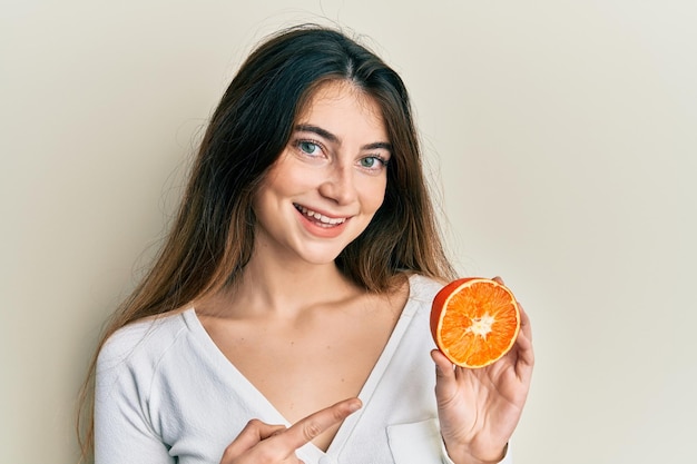 Junge kaukasische Frau hält eine frische Orangenscheibe in der Hand, lächelt glücklich und zeigt mit Hand und Finger