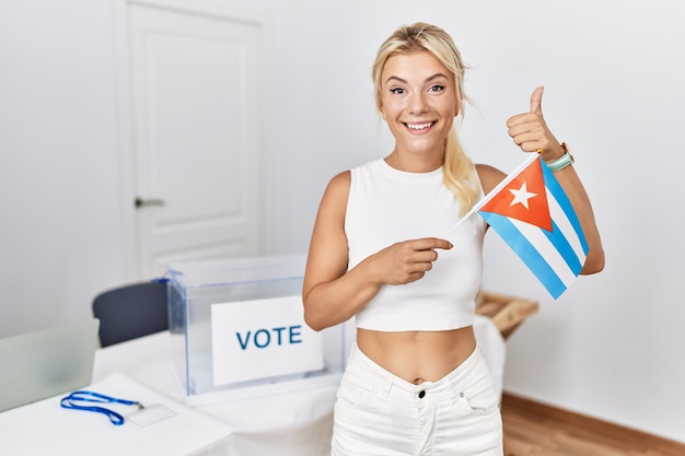 Junge kaukasische Frau im politischen Wahlkampf hält die kubanische Flagge in der Hand, lächelt glücklich und positiv, zeigt den Daumen nach oben und macht ein hervorragendes und positives Zeichen