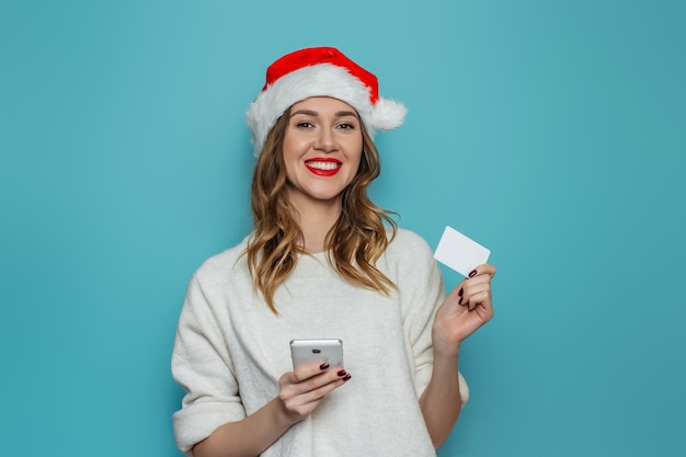 Junge kaukasische Frau im Weihnachtsmannhut und im weißen Winterpullover lächelnd, Handy und Kreditkarte haltend